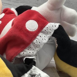 May include: A Minnie Mouse soft toy, dressed in a red dress with white polka dots and white lace trim. The toy has white gloves, black shoes, and a yellow skirt. The Disney tag is visible.