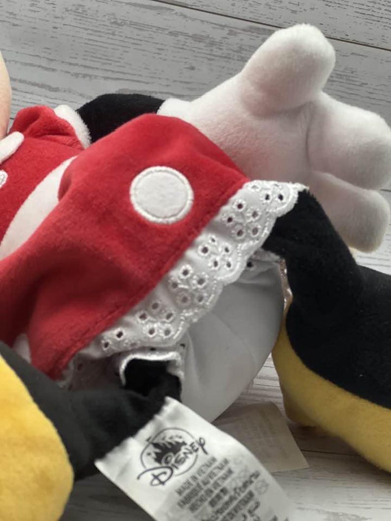 May include: A Minnie Mouse soft toy, dressed in a red dress with white polka dots and white lace trim. The toy has white gloves, black shoes, and a yellow skirt. The Disney tag is visible.