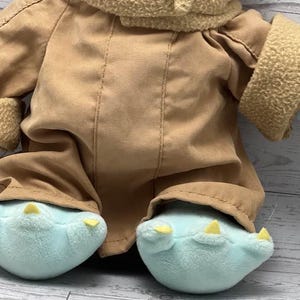 May include: A plush doll with a tan coat and light blue feet with yellow claws. The doll has a tan, textured collar and sleeves. The doll's outfit is a soft, brown fabric. The doll is a character from a popular science fiction franchise.