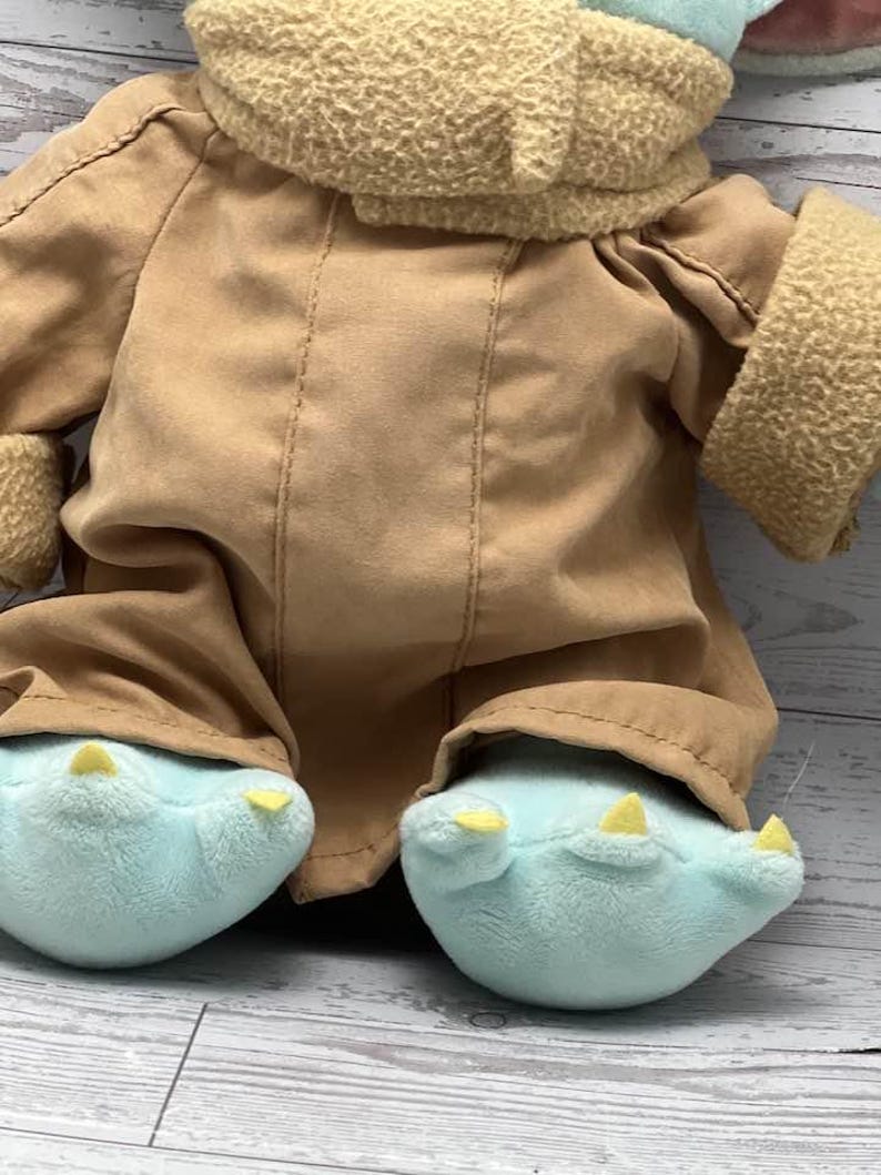 May include: A plush doll with a tan coat and light blue feet with yellow claws. The doll has a tan, textured collar and sleeves. The doll's outfit is a soft, brown fabric. The doll is a character from a popular science fiction franchise.