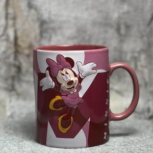 Op de afbeelding: Een keramische mok met een afbeelding van Minnie Mouse in een roze jurk met strik. De mok heeft een roze handvat en binnenkant, met een achtergrond van overlappende roze en donkerrode geometrische vormen en de tekst MINNIE in het wit.