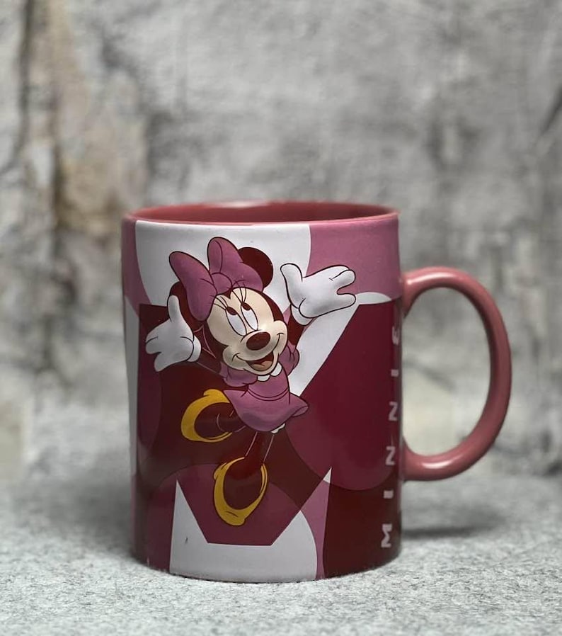 Op de afbeelding: Een keramische mok met een afbeelding van Minnie Mouse in een roze jurk met strik. De mok heeft een roze handvat en binnenkant, met een achtergrond van overlappende roze en donkerrode geometrische vormen en de tekst MINNIE in het wit.