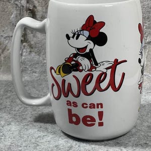 Pu&ograve; includere: Una tazza in ceramica bianca con un'illustrazione di Minnie Mouse che indossa un fiocco rosso e un vestito a pois. Il testo "Sweet as can be!" &egrave; stampato in rosso, in corsivo e in stampatello, sotto il personaggio. La tazza ha un manico curvo.