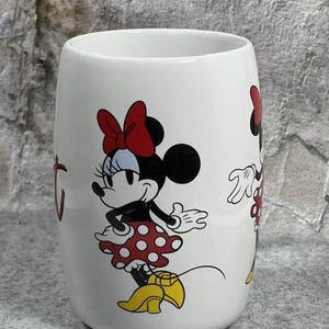 Pu&ograve; includere: Una tazza in ceramica bianca con una grafica di Minnie Mouse che indossa una gonna a pois rossi, un fiocco rosso e scarpe gialle. Il personaggio &egrave; raffigurato in piedi con una mano sul fianco. Il disegno &egrave; stampato sulla superficie esterna.