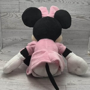 May include: A plush Minnie Mouse doll, viewed from the back, with black ears, a pink bow, and a pink dress. The doll has white gloves and black and white sleeves. A black tail extends from the back of the doll.