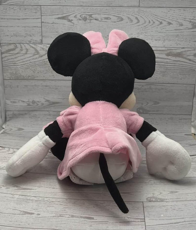 May include: A plush Minnie Mouse doll, viewed from the back, with black ears, a pink bow, and a pink dress. The doll has white gloves and black and white sleeves. A black tail extends from the back of the doll.