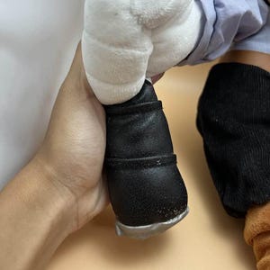 May include: A plush toy's leg and foot are visible, featuring a black shoe with a silver sole. The leg is covered in a light blue fabric and a white plush hand. The toy's design suggests a cartoon character.