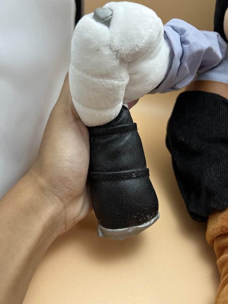 May include: A plush toy's leg and foot are visible, featuring a black shoe with a silver sole. The leg is covered in a light blue fabric and a white plush hand. The toy's design suggests a cartoon character.