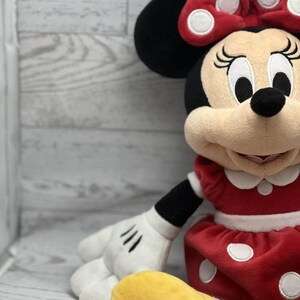 May include: A plush Minnie Mouse doll. The doll has a red dress with white polka dots, a matching bow, and black ears. The doll has a cream-coloured face, black eyes, and a smiling expression. The doll's hands are white with black gloves.