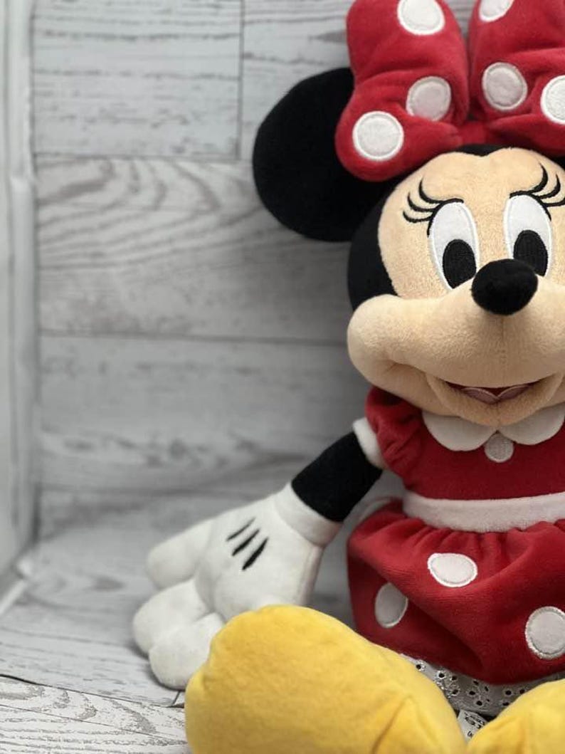 May include: A plush Minnie Mouse doll. The doll has a red dress with white polka dots, a matching bow, and black ears. The doll has a cream-coloured face, black eyes, and a smiling expression. The doll's hands are white with black gloves.