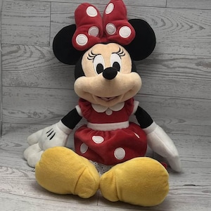 May include: A Minnie Mouse soft toy. The doll features a red dress with white polka dots, a matching bow, and yellow shoes. Minnie's face is beige with black eyes and a black nose. The doll has black ears and arms, and white gloves.