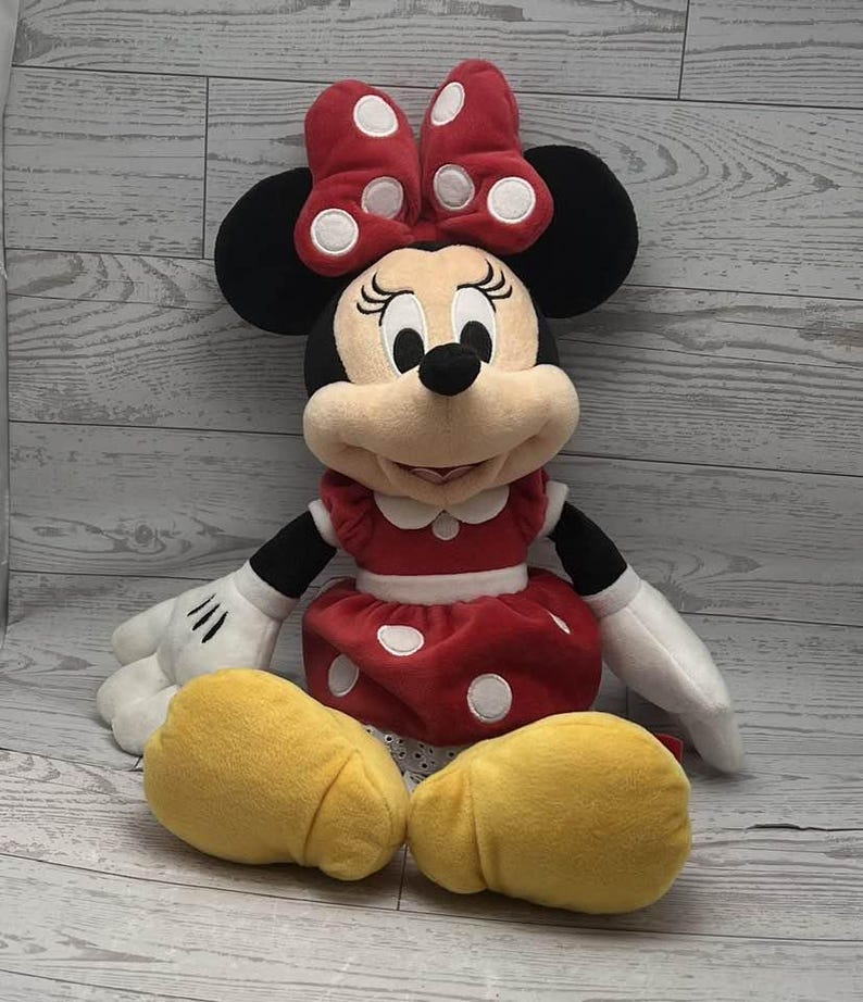 May include: A Minnie Mouse soft toy. The doll features a red dress with white polka dots, a matching bow, and yellow shoes. Minnie's face is beige with black eyes and a black nose. The doll has black ears and arms, and white gloves.