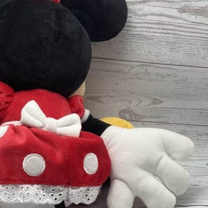 May include: A plush Minnie Mouse doll with a black head and large ears, a red dress with white polka dots, and a white bow. The doll has white gloves and a black tail. The doll is made of soft, plush material.