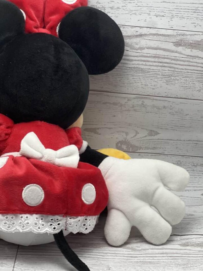 May include: A plush Minnie Mouse doll with a black head and large ears, a red dress with white polka dots, and a white bow. The doll has white gloves and a black tail. The doll is made of soft, plush material.