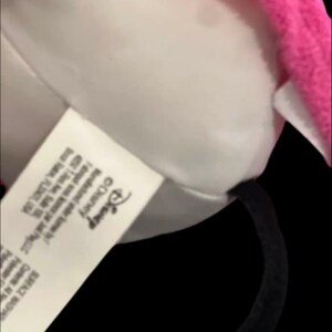 May include: A close-up of a plush Minnie Mouse ear headband. The headband features a white plush ear with a pink trim. A tag with the Disney logo is visible.