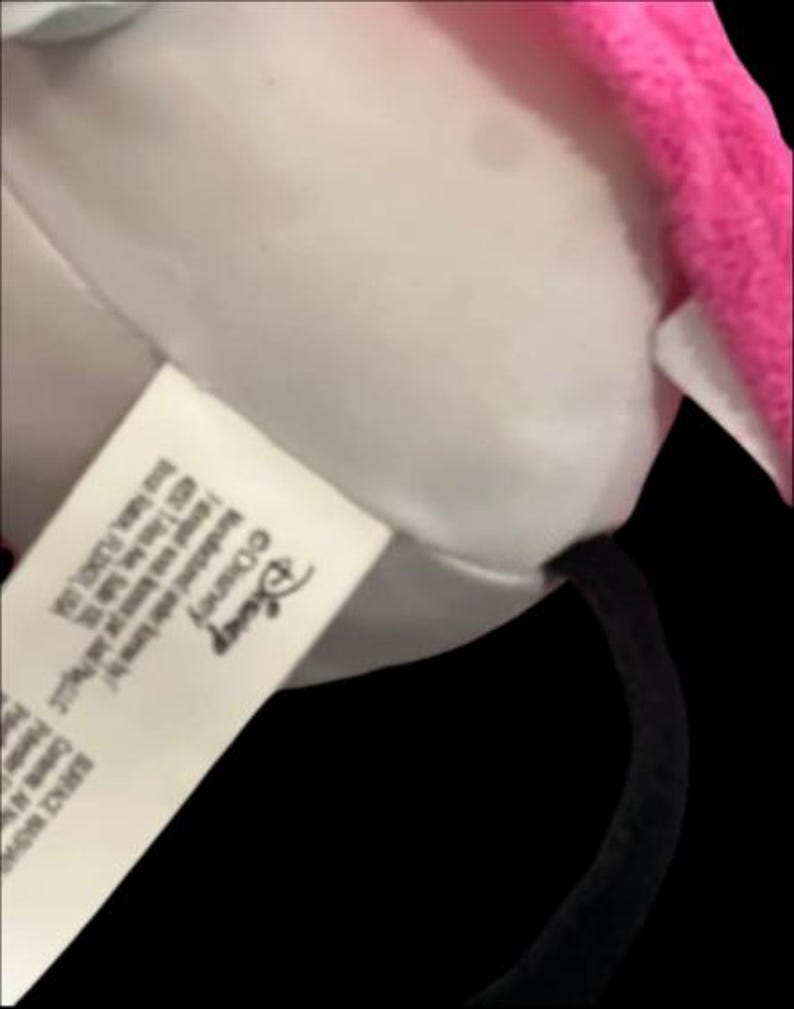 May include: A close-up of a plush Minnie Mouse ear headband. The headband features a white plush ear with a pink trim. A tag with the Disney logo is visible.