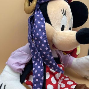 May include: A plush Minnie Mouse doll, featuring a black head with large ears, a beige face, and a red and white polka-dot dress. The doll wears a purple scarf with white polka dots and a red hat with yellow accents.
