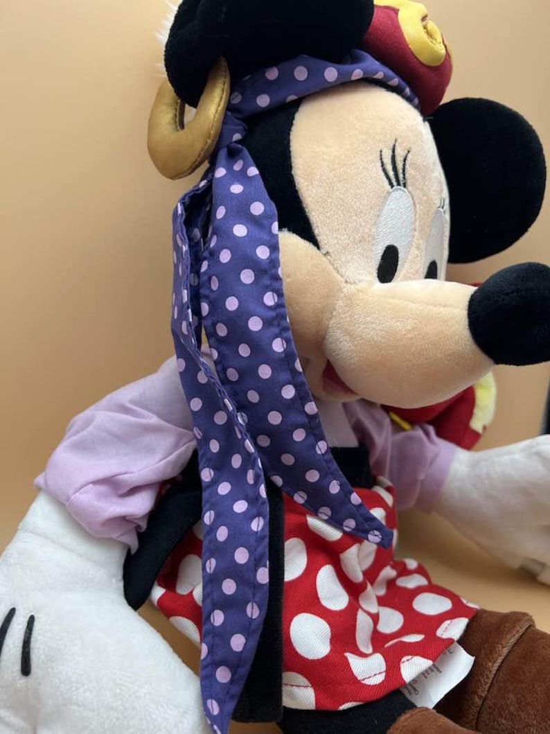 May include: A plush Minnie Mouse doll, featuring a black head with large ears, a beige face, and a red and white polka-dot dress. The doll wears a purple scarf with white polka dots and a red hat with yellow accents.