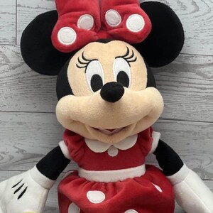 May include: A plush Minnie Mouse doll in a red dress and bow, both with white polka dots. The doll has black ears, a black nose, and white gloves. The background is a light wood grain.
