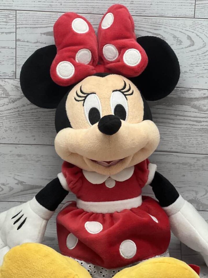 May include: A plush Minnie Mouse doll in a red dress and bow, both with white polka dots. The doll has black ears, a black nose, and white gloves. The background is a light wood grain.