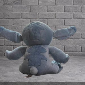 Puede incluir: Un peluche azul de un personaje alien&iacute;gena con grandes orejas y brazos extendidos. El juguete est&aacute; sentado sobre una superficie gris frente a una pared de ladrillos grises.