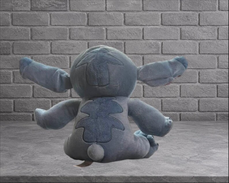 Puede incluir: Un peluche azul de un personaje alien&iacute;gena con grandes orejas y brazos extendidos. El juguete est&aacute; sentado sobre una superficie gris frente a una pared de ladrillos grises.