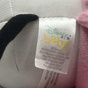 May include: A close-up of a Disney Baby plush toy with a tag. The tag features the Disney Baby logo in yellow and the text "&copy; Disney". The plush toy is white, black, and pink.