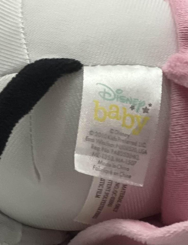 May include: A close-up of a Disney Baby plush toy with a tag. The tag features the Disney Baby logo in yellow and the text "&copy; Disney". The plush toy is white, black, and pink.