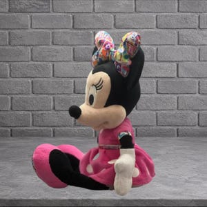 May include: A plush Minnie Mouse doll, seated with a pink dress and bow. The doll has a black head, ears, and tail, with a beige face and a black nose. The bow is multi-colored. The dress has white polka dots.