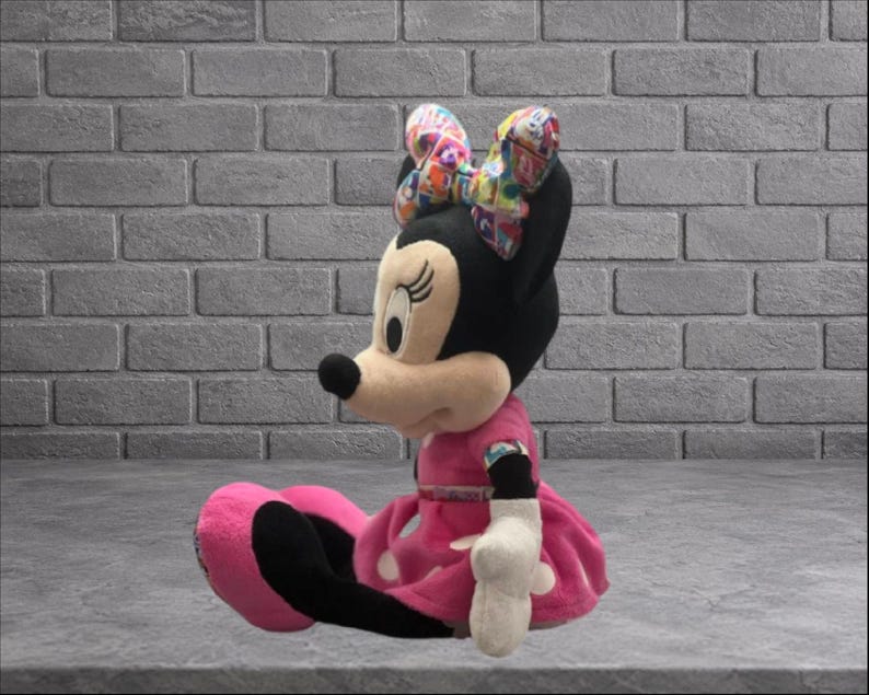 May include: A plush Minnie Mouse doll, seated with a pink dress and bow. The doll has a black head, ears, and tail, with a beige face and a black nose. The bow is multi-colored. The dress has white polka dots.