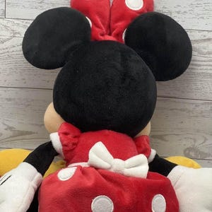 May include: A plush Minnie Mouse doll, seen from the back, with black ears, a red bow with white spots, and a red dress with white spots and a white bow. The doll has white gloves and yellow shoes.