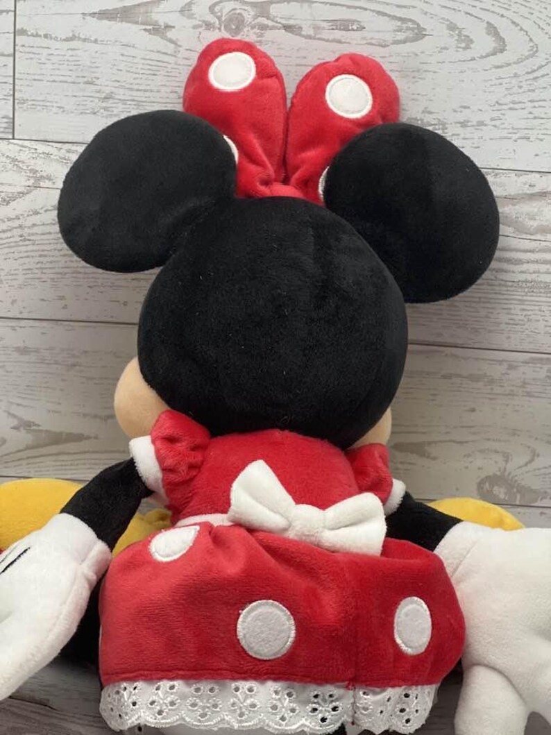 May include: A plush Minnie Mouse doll, seen from the back, with black ears, a red bow with white spots, and a red dress with white spots and a white bow. The doll has white gloves and yellow shoes.