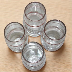 May include: Four clear, stemless drinking glasses with a rounded shape. Each glass has a thin, dark red band near the rim and a faint design with the word "final". The glasses are arranged on a light brown wooden surface.