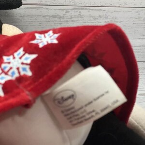 Puede incluir: Un peluche con un gorro rojo adornado con dise&ntilde;os de copos de nieve blancos y azules. El gorro est&aacute; sobre un peluche blanco y negro. El logotipo de Disney es visible en una etiqueta.