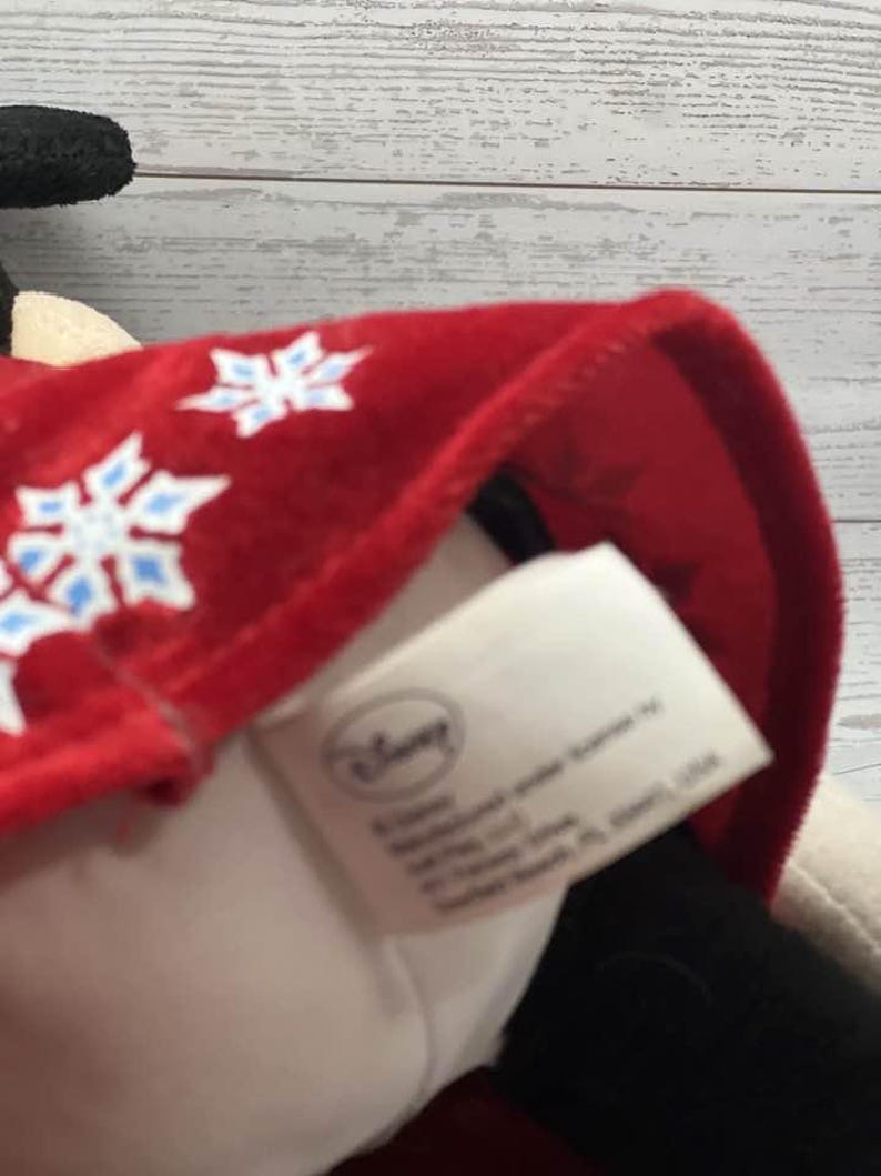 Puede incluir: Un peluche con un gorro rojo adornado con dise&ntilde;os de copos de nieve blancos y azules. El gorro est&aacute; sobre un peluche blanco y negro. El logotipo de Disney es visible en una etiqueta.