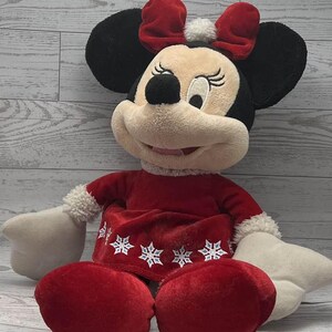 Puede incluir: Un mu&ntilde;eco de peluche de Minnie Mouse con un atuendo festivo. El mu&ntilde;eco tiene orejas negras, un lazo rojo y un vestido rojo con detalles de copos de nieve. Las manos y los pies del mu&ntilde;eco son blancos.