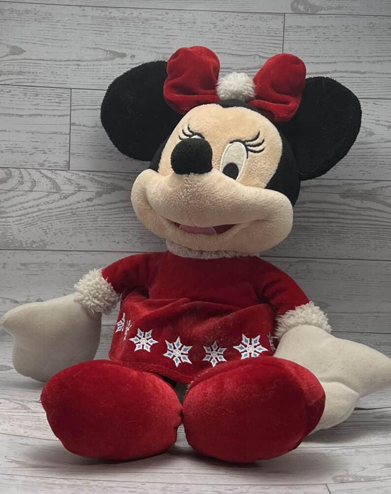 Puede incluir: Un mu&ntilde;eco de peluche de Minnie Mouse con un atuendo festivo. El mu&ntilde;eco tiene orejas negras, un lazo rojo y un vestido rojo con detalles de copos de nieve. Las manos y los pies del mu&ntilde;eco son blancos.
