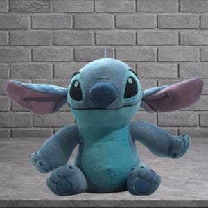 Puede incluir: Un peluche del personaje Stitch de la pel&iacute;cula de Disney Lilo & Stitch. El juguete es principalmente azul con orejas grandes, ojos negros y una peque&ntilde;a antena. Est&aacute; sentado frente a una pared de ladrillos.