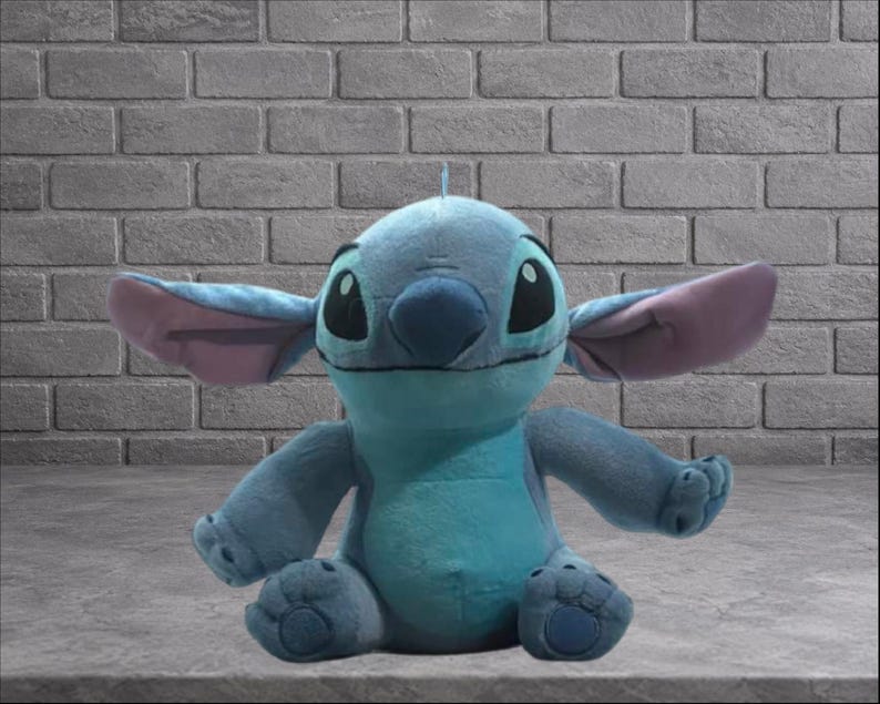 Puede incluir: Un peluche del personaje Stitch de la pel&iacute;cula de Disney Lilo & Stitch. El juguete es principalmente azul con orejas grandes, ojos negros y una peque&ntilde;a antena. Est&aacute; sentado frente a una pared de ladrillos.