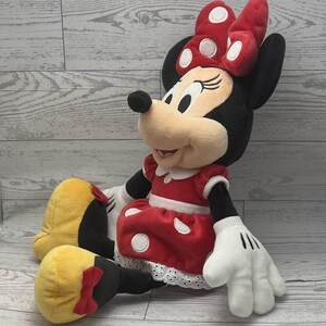 May include: A plush Minnie Mouse doll. Minnie is wearing a red dress with white polka dots, a matching bow, and white gloves. Her yellow shoes have red bows. The doll is sitting on a light wood-grain surface.