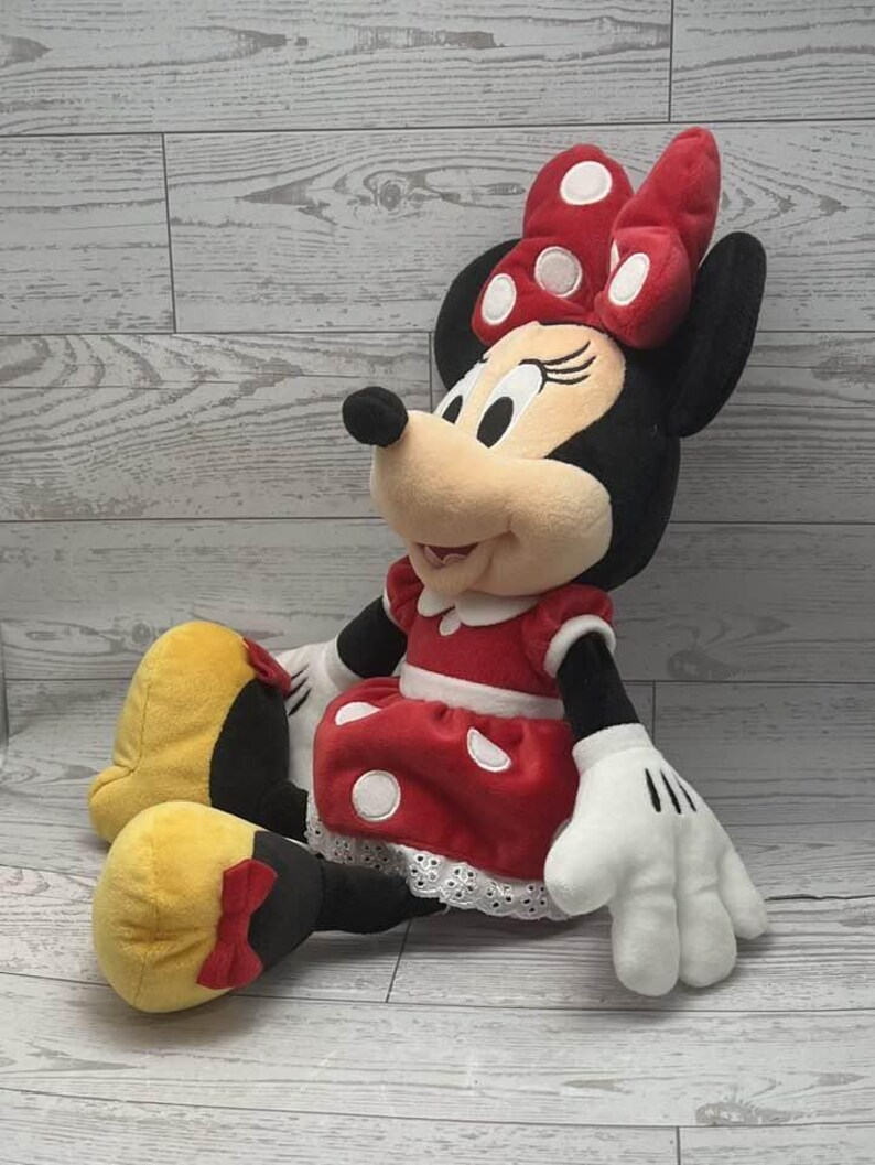 May include: A plush Minnie Mouse doll. Minnie is wearing a red dress with white polka dots, a matching bow, and white gloves. Her yellow shoes have red bows. The doll is sitting on a light wood-grain surface.