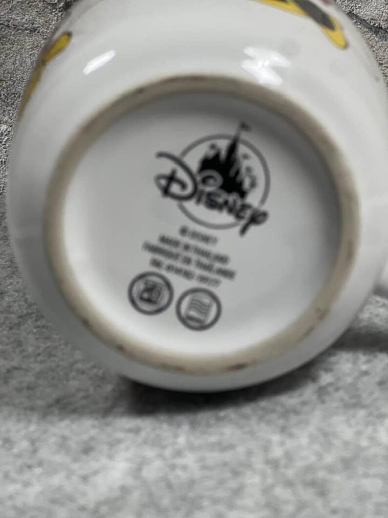 Pu&ograve; includere: Il fondo di una tazza in ceramica bianca con un logo Disney nero e la sagoma di un castello. Il testo include Disney, Made in Thailand e Fabriqu&eacute; en Tha&iuml;lande. Sono presenti due icone circolari che raffigurano un microonde e una lavastoviglie.