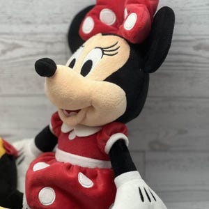 May include: A Minnie Mouse soft toy, dressed in a red dress with white polka dots, a matching bow, and white gloves. The doll has black ears, a black nose, and a cream-coloured face. A classic character.
