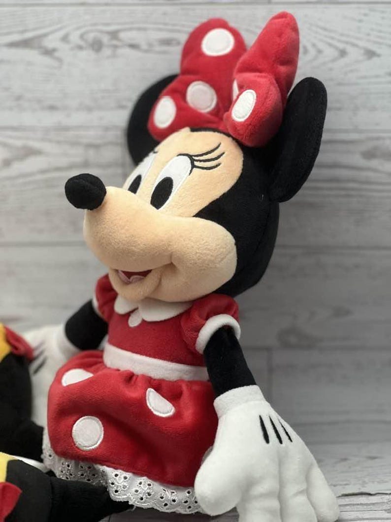 May include: A Minnie Mouse soft toy, dressed in a red dress with white polka dots, a matching bow, and white gloves. The doll has black ears, a black nose, and a cream-coloured face. A classic character.