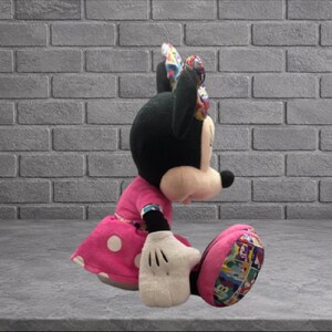 May include: A plush Minnie Mouse doll in a seated position. The doll has a black head and ears, a pink dress with white polka dots, and white gloves. The bow and shoe details feature a colorful pattern.