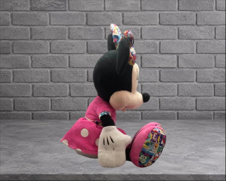 May include: A plush Minnie Mouse doll in a seated position. The doll has a black head and ears, a pink dress with white polka dots, and white gloves. The bow and shoe details feature a colorful pattern.