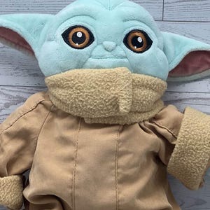 May include: A soft toy of a character with big brown eyes, large ears, and a light blue head. The doll is wearing a tan coat and a beige scarf. The toy is a character from a popular science fiction franchise.