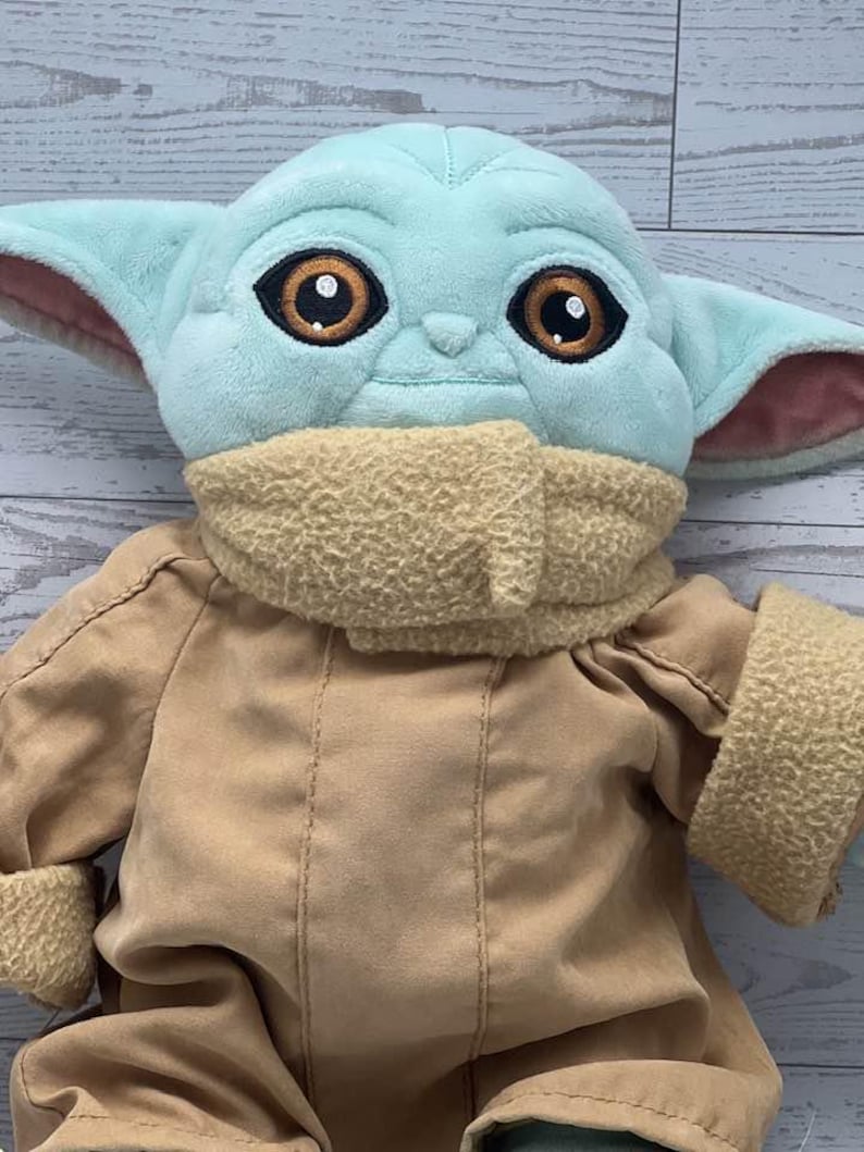 May include: A soft toy of a character with big brown eyes, large ears, and a light blue head. The doll is wearing a tan coat and a beige scarf. The toy is a character from a popular science fiction franchise.