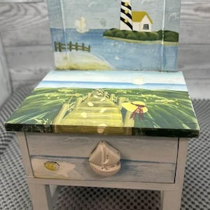 May include: A small, hand-painted wooden chair with a drawer, featuring a coastal theme. The back of the chair depicts a lighthouse and ocean scene. The drawer has a sailboat-shaped handle and a painted landscape on top. The chair is painted in shades of blue, white, and green.