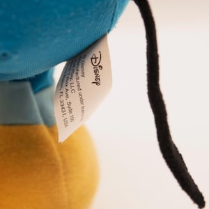 May include: A plush toy featuring a bright blue fabric section, a yellow fabric base, and a thin black tail. A white fabric tag is attached, displaying the Disney logo and text: Manufactured under license by Just Play, LLC, 4850 T-Rex Ave, Suite 100, Boca Raton, FL 33431, USA.